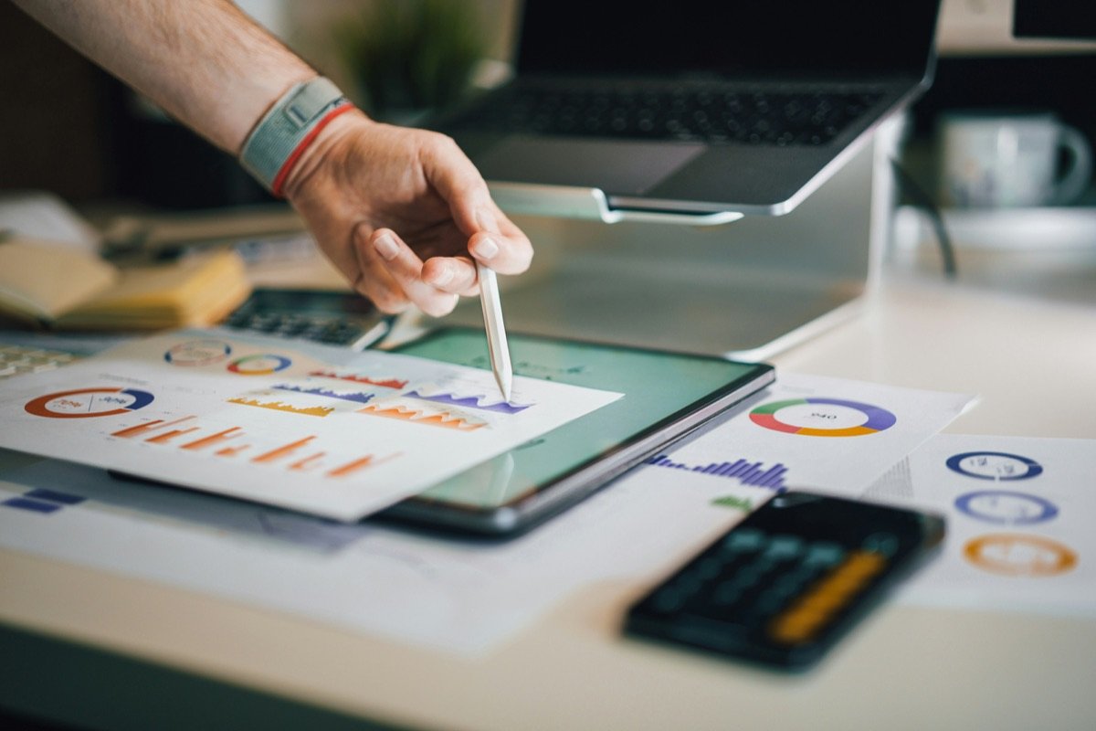 A hand with a stylus over a tablet and printed charts